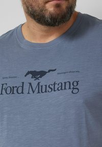 Jasnoniebieska koszulka z miękkiego materiału, z czarnym sylwetką konia Mustang oraz napisem "Ford Mustang" i "Sportowy, luksusowy ...".