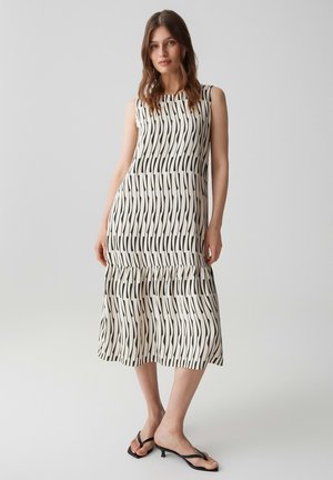 Femme portant une robe blanche sans manches, mi-longue, avec des rayures verticales ondulées noires et des sandales noires à talons bas, debout devant un fond uni.