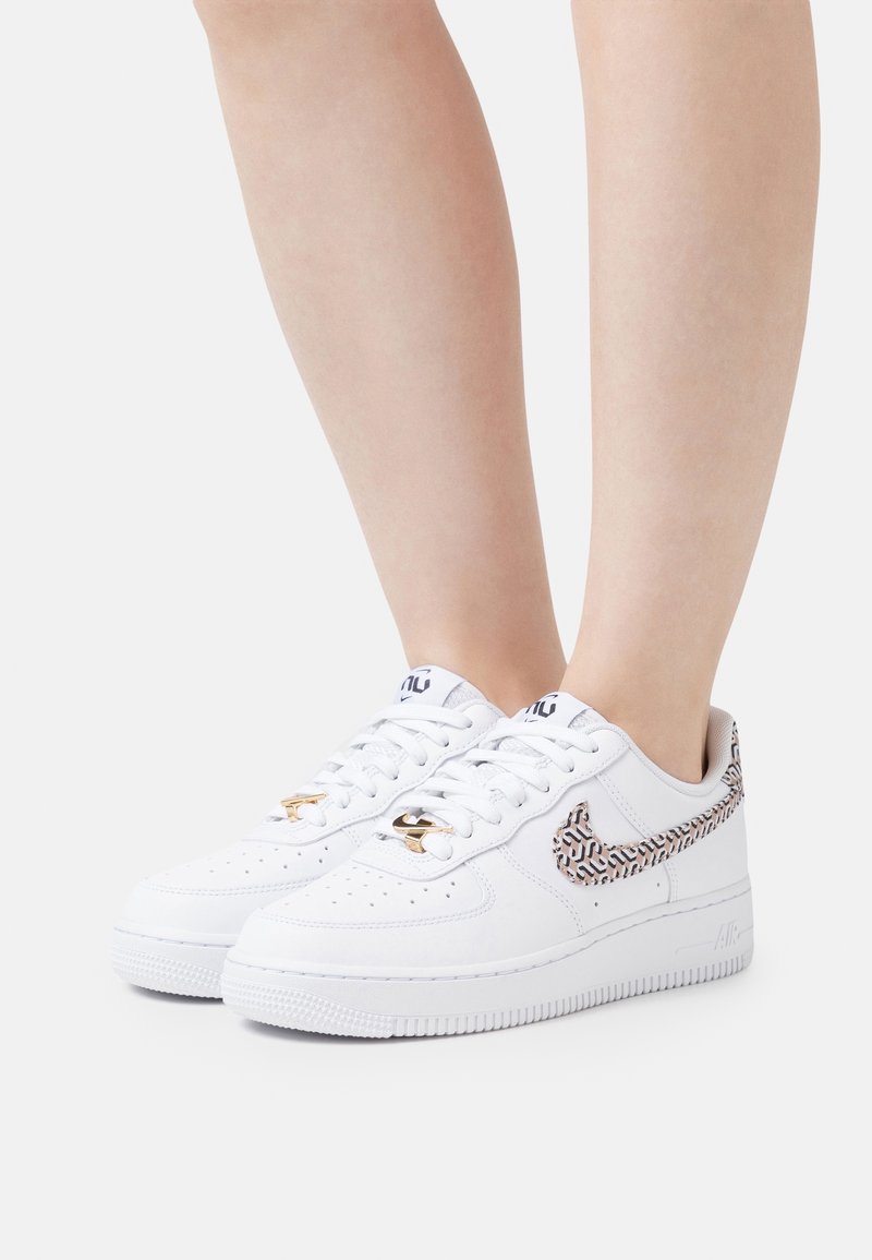 Baskets basses blanches avec swoosh Nike à motif et breloques dorées aux lacets, portées pieds nus sur un fond uni.