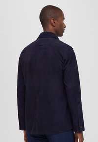 Chaqueta de ante azul marino con cuello clásico, mangas largas y puños abotonados. Silueta limpia que presenta un detalle de costura central en la parte posterior.