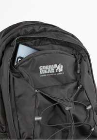 Black backpack featuring a front zip pocket with an interior device, textured fabric, adjustable bungee cords, and a logo printed inside.