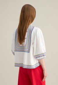 Blusa blanca con bordado colorido en el cuello y las mangas, con mangas cortas abullonadas y un corte holgado. Detalles de costura visibles.