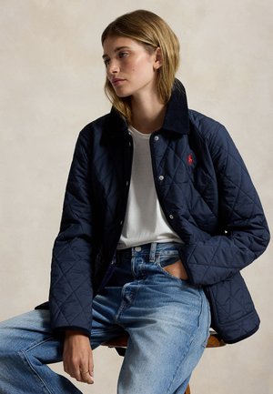 Jeune femme assise sur un tabouret, portant une veste matelassée bleu marine, un t-shirt blanc et un jean bleu clair, regardant sur le côté.