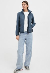 Tumesin fleece jakk kõrge kraega, lukuga ja kahe küljetaskuga. Omab kontrastset tumedat varrukat ja heledat tekstuuri. Kombineeritud heledate teksadega.