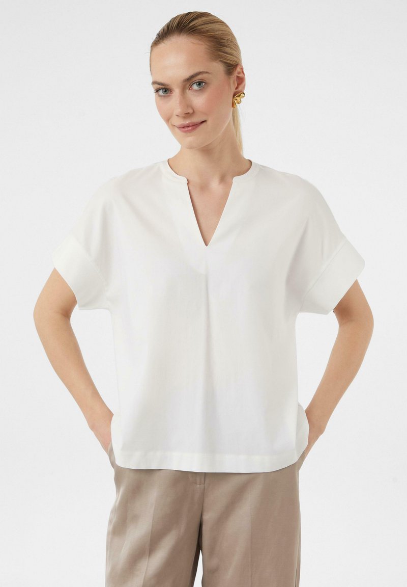 Weiße Bluse mit kurzen Ärmeln und V-Ausschnitt, mit umgeschlagenen Bündchen, aus glattem Stoff, kombiniert mit beigen Hosen.