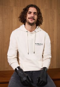 Krēmas krāsas kapuce ar priekšējo logotipu, auklām un ribotām aprocēm, apvienota ar melnām cimdiem un tumšām džinsiem, uz koka fona.