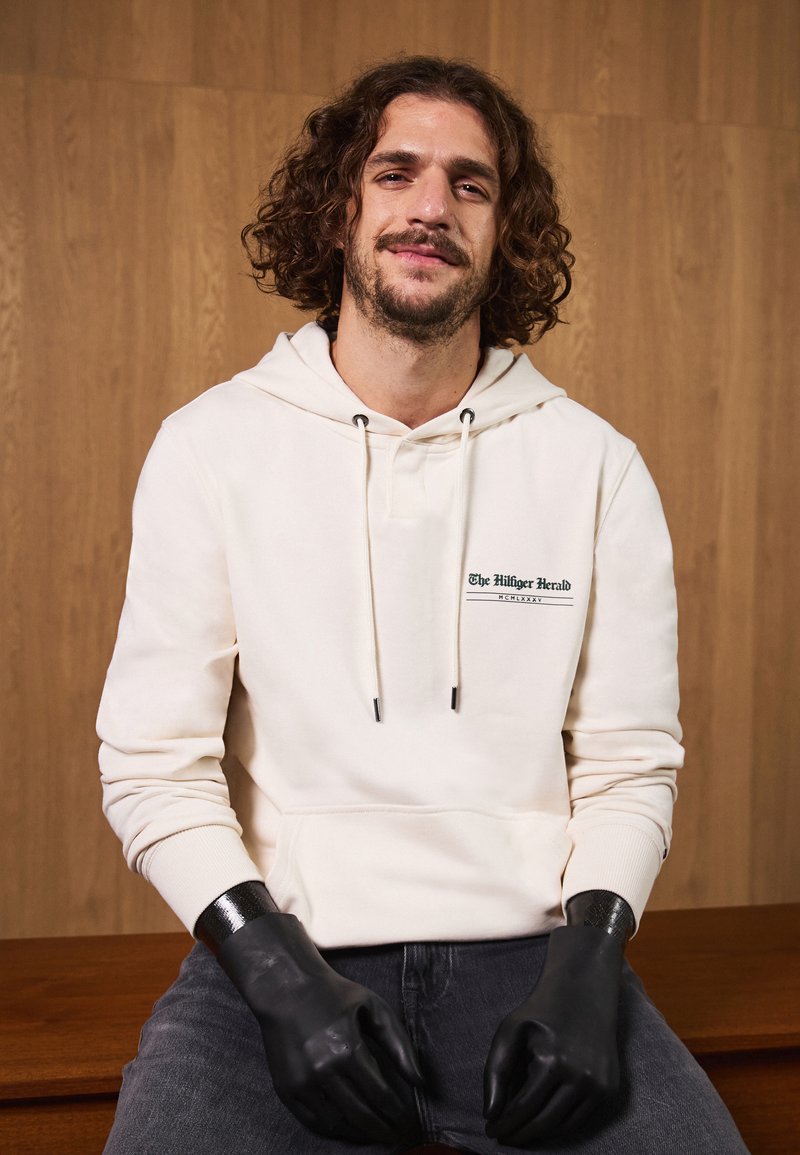 Krēmas krāsas kapuce ar priekšējo logotipu, auklām un ribotām aprocēm, apvienota ar melnām cimdiem un tumšām džinsiem, uz koka fona.