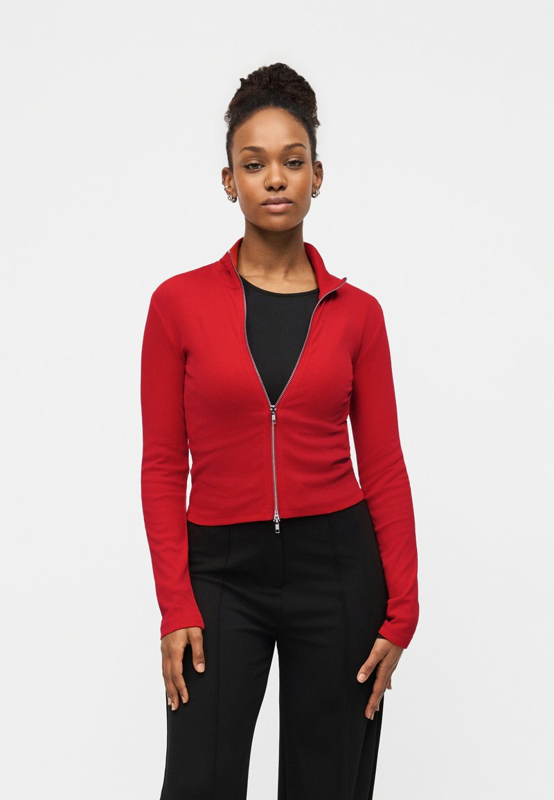 Junge Frau mit zusammengebundenen Haaren, die eine rote Jacke mit Reißverschluss über einem schwarzen Shirt und schwarze Hosen trägt, vor einem weißen Hintergrund stehend.