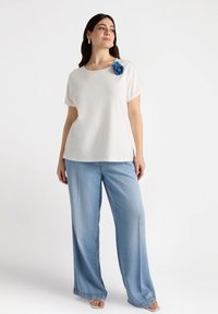 Top blanco de manga corta con un detalle de flor de tela azul, combinado con jeans amplios de un azul claro. Textura de algodón suave con un ajuste relajado.