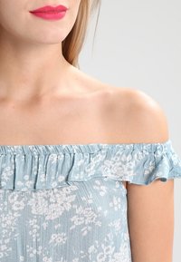 Světle modrý top bez ramínek s volánkovým výstřihem, květinový vzor v bílé barvě, vyrobený z měkké tkaniny, s texturovaným povrchem.