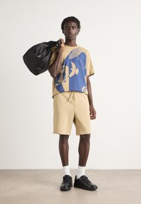 T-shirt beige surdimensionnée avec un graphique bleu, associée à un short beige et des baskets noires. Le modèle porte un sac noir et se tient sur un sol lisse.