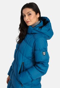 Gewatteerde blauwe jas met hoge kraag, afneembare capuchon, zijzakken en logo-opdruk op de linker mouw. Glad, glanzend materiaal.