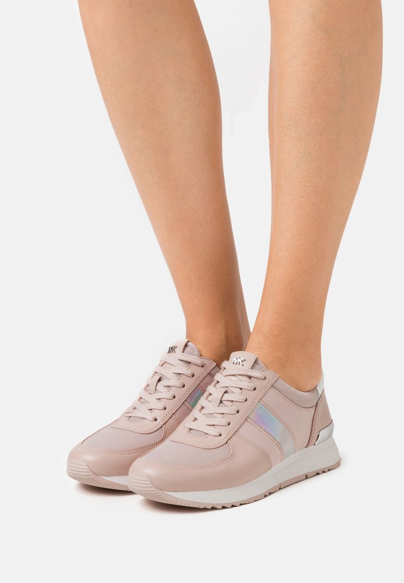 Chaussures de sport rose clair avec une texture lisse, en mesh et matériaux synthétiques, une bande latérale irisée et une semelle en caoutchouc blanche.
