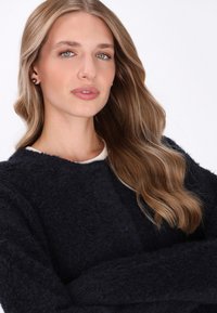 Blonde vrouw met golvend haar draagt een zwarte, fuzzy, knooptrui en pareloorbellen, kijkt zelfverzekerd naar de camera met gekruiste armen.