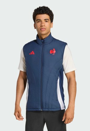 Navy padded vest with high collar, front zipper, red accents (logo and stripes) and side pockets, worn over a light-colored shirt.