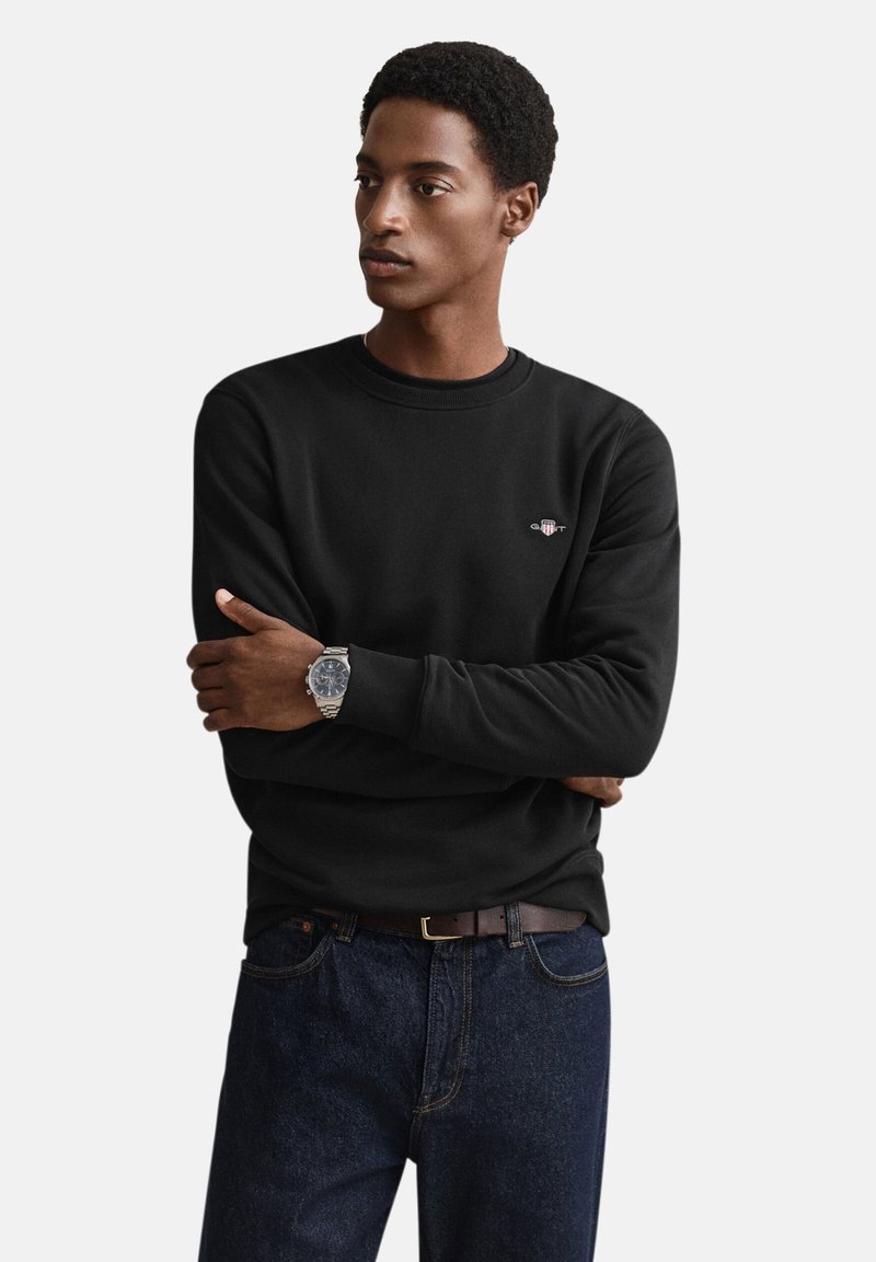 Zwarte katoenen sweatshirt met ronde hals, lange mouwen en een klein logo op de borst, gecombineerd met donkerdenim jeans en een zilveren horloge.