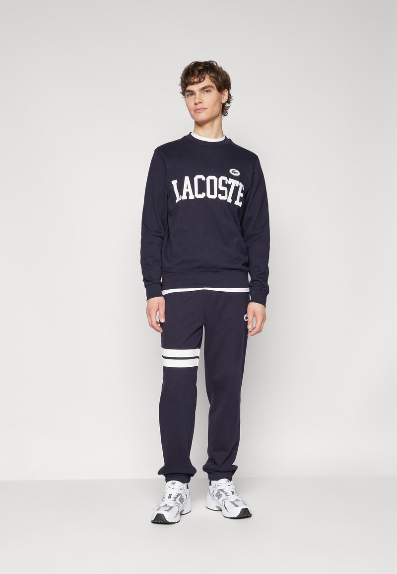 Sudadera azul marina con el logo blanco "LACOSTE", combinada con pantalones a juego que presentan rayas blancas y logos. El material parece suave y liso.
