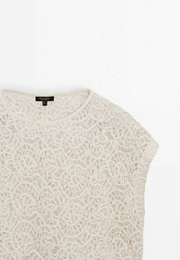 Top en dentelle blanc cassé avec un motif floral, décolleté rond, manches courtes et une finition douce et texturée. Inclut une fermeture au dos.