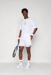 Camiseta blanca de Nike con logo dorado, pantalones cortos blancos y zapatillas blancas. Sostiene una raqueta de tenis y una pelota, con muñequeras y cabello oscuro.