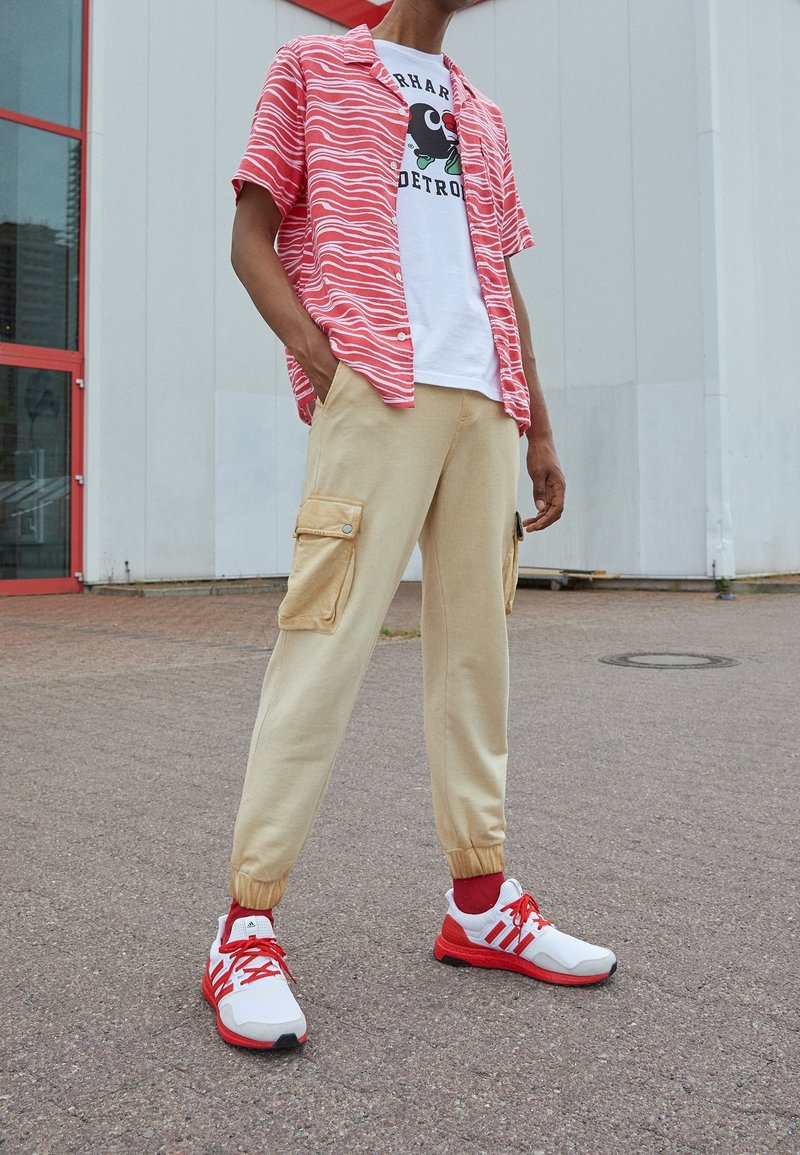 Růžová pruhovaná košile s krátkým rukávem, bílý tričko s grafikou, béžové tepláky s cargo kapsami a červenobílé tenisky.
