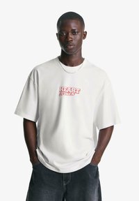 Tricou alb din bumbac cu text roșu "HEART struck", mâneci scurte, croială lejeră și decolteu rotund. Asortat cu pantaloni scurți din denim închis.