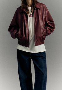Burgundowa skórzana kurtka bomberka z elastycznymi mankietami, zamek błyskawiczny i boczne kieszenie, noszona na białej koszulce i ciemnoniebieskich dżinsach.