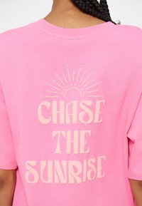 Човек с плитка, носещ розова риза с текст "CHASE THE SUNRISE" и частична графика на изгрев отзад.