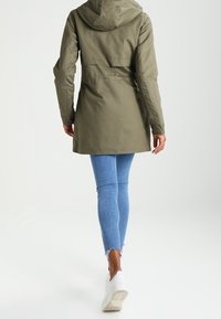 Zelená nepromokavá parka s kapucí, střední délky, se staženým pasem, kombinovaná se světle modrými skinny džíny a bílými teniskami.