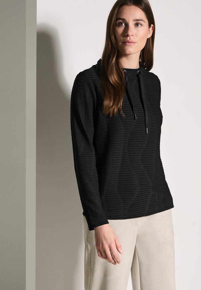 Schwarzer strukturierter Pullover mit langen Ärmeln und Kapuze, ausgestattet mit einem Wellenmuster. Kombiniert mit hellen, weit geschnittenen Hosen.