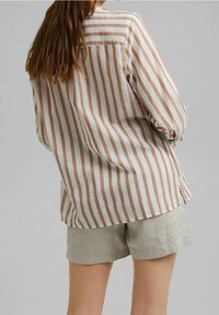 Chemise rayée à manches longues en crème et marron clair, avec une coupe ample, des poignets boutonnés et des fentes sur les côtés, portée avec un short gris clair.