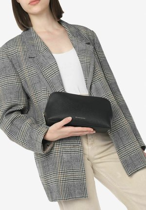 Sort læderpouch med en buet top og guldpræget logo. Holdt i en hånd, sat mod en gråternet blazer og lyse bukser.