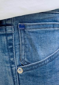 Plave traperice s ušivenim džepom s metalnom zakovicom i plavom etiketom označenom "J&J 1990." Tekstura je meka s izblijedjelim izgledom.
