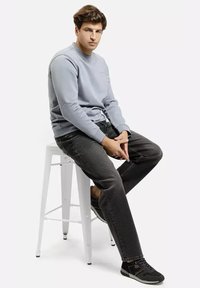 Jasnoniebieska bluza z okrągłym dekoltem, w zestawieniu z ciemnoszarymi dżinsami. Model siedzi na białym hokerze, nosząc czarne buty z czerwonymi akcentami.