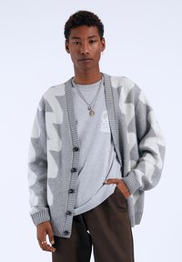 Gray and white patterned cardigan with a button closure and ribbed cuffs, layered over a light gray shirt, worn with brown pants.