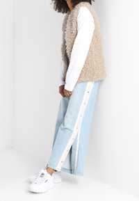 Beige faux fur väst, vit långärmad skjorta, ljusblå vida jeans med sidoknappar och vita sneakers. Enkla, mjuka texturer.