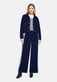 Granatowa kurtka z dwoma przednimi kieszeniami i zapięciem na guziki, zestawiona z granatowymi spodniami o szerokich nogawkach, ozdobionymi jasnoniebieskimi i kremowymi paskami po bokach.