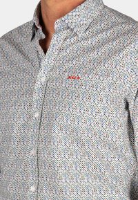 Overhemd met knopen op een witte achtergrond, met een meerkleurig stippenpatroon in blauw, groen, rood en geel, en een rood logo op de borst.