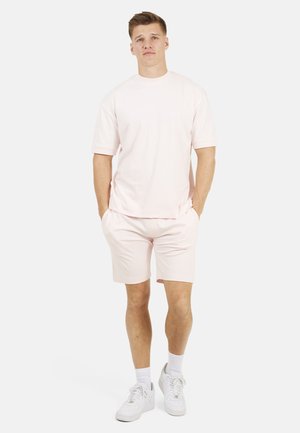 Młody mężczyzna stojący z rękami w kieszeniach, ubrany w jasnoróżowy T-shirt, pasujące szorty, białe skarpetki i białe trampki na białym tle.