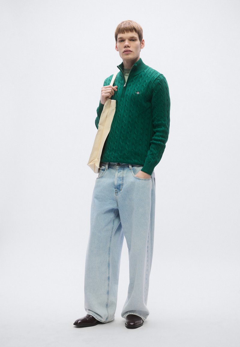 Green cable-knit sweater with a half-zip, paired with light blue oversized jeans and dark shoes. Model holds a beige tote bag.