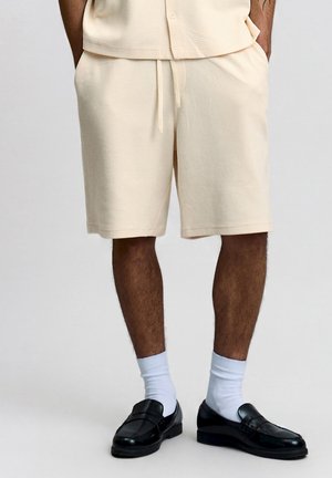 Mand iført cremet strukturerede shorts, hvide sokker og sorte læder loafers står med hænderne i lommerne.