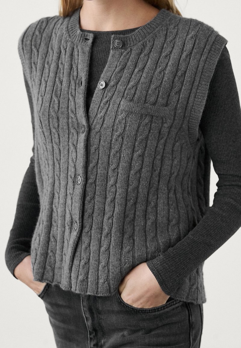 Graue Strickweste mit Rundhalsausschnitt, Knöpfen vorne und einer aufgesetzten Tasche; getragen über einem dunkelgrauen Langarmshirt.