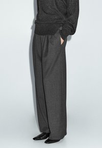 Graue weit geschnittene Hosen aus strukturiertem Stoff, mit tiefen Taschen, hoher Taille und lockerer Passform, kombiniert mit spitzen schwarzen Schuhen.