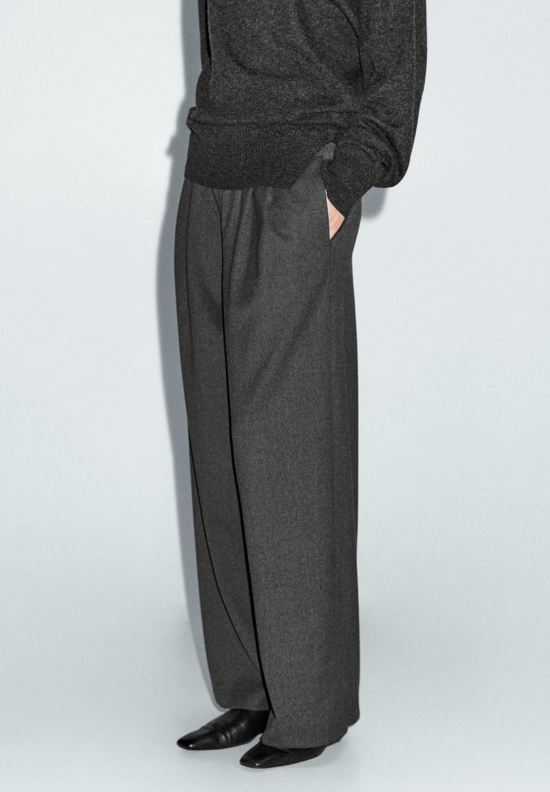 Graue weit geschnittene Hosen aus strukturiertem Stoff, mit tiefen Taschen, hoher Taille und lockerer Passform, kombiniert mit spitzen schwarzen Schuhen.