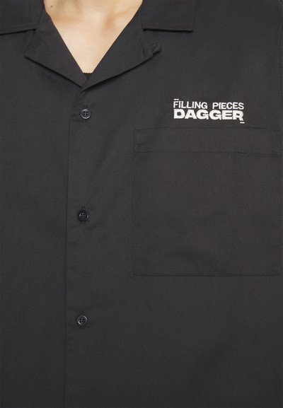 Melnas pogājamās krekls ar krūšu kabatu, kurā ir balts izšūts teksts "Filling Pieces DAGGER" augšējā kreisajā pusē.