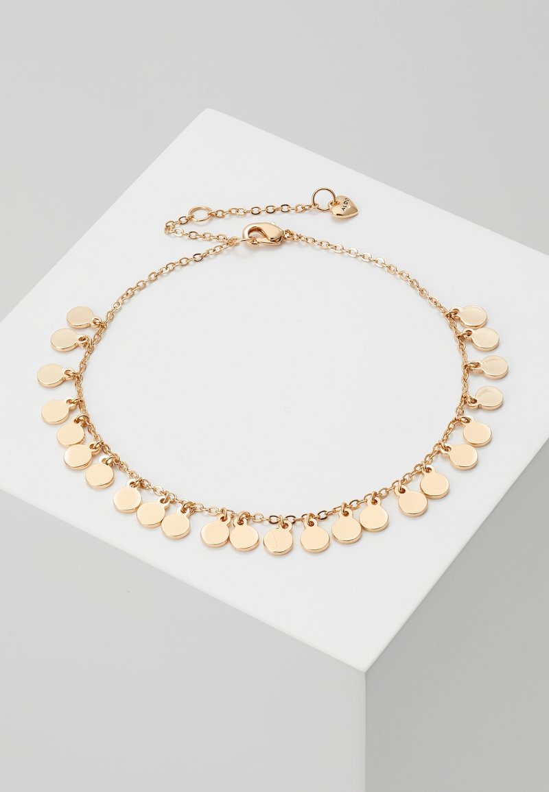 Bracelet chaîne en or avec plusieurs petits charms ronds en forme de disque et une petite étiquette en forme de cœur sur un bloc carré blanc.