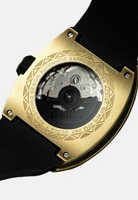 Goudkleurige automatische achterzijde van het horloge met ingewikkelde gravures, die tandwielen toont en een zwarte rubberen band heeft. Saffierglas, waterbestendig tot 50 meter.