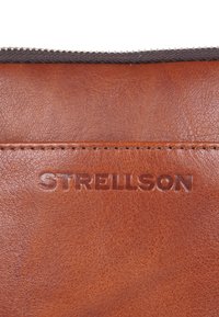 Borsa in pelle marrone con finitura testurizzata e logo "STRELLSON" in rilievo. Presenta una zip grigia a contrasto nella parte superiore.