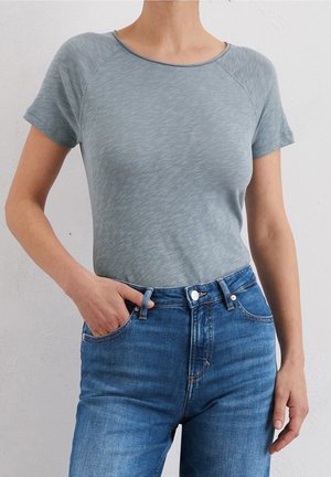 Vrouw draagt een aangesloten lichtgrijs shirt met korte mouwen en een hooggetailleerde blauwe spijkerbroek, met één hand in een zak, tegen een effen witte muur.