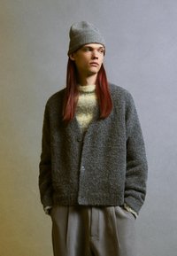 Cardigan grigio con texture, a V, con bottoni frontali, indossato sopra un maglione a righe gialle e grigie, abbinato a pantaloni grigio chiaro e un berretto in maglia.