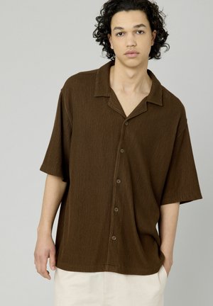 Chemise brune à rayures avec des manches courtes, un col pointu et cinq boutons sur le devant, portée sur un short de couleur crème.